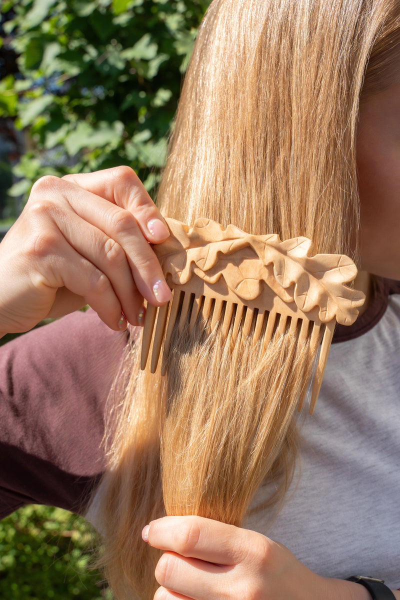 A close-up lifestyle shot of a person holding a long, light brown wooden hair comb through their blonde hair outdoors, with green foliage blurred in the background.
