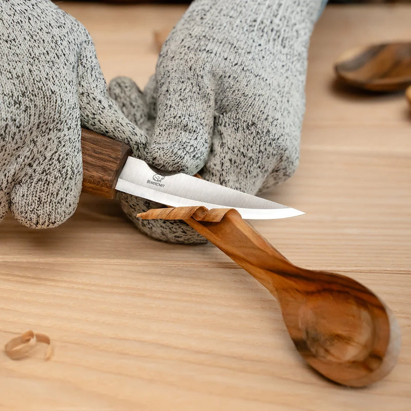 Shaping a wooden spoon with a carving knife, hands in cut-resistant gloves