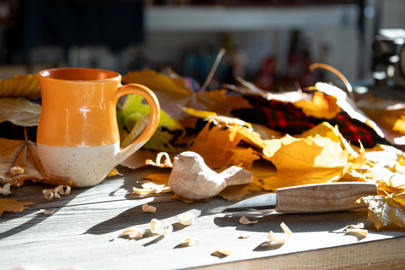An orange and white ceramic mug sits on a rustic wooden table surrounded by scattered autumn leaves and wood shavings, with a knife nearby in a warm, natural setting.
