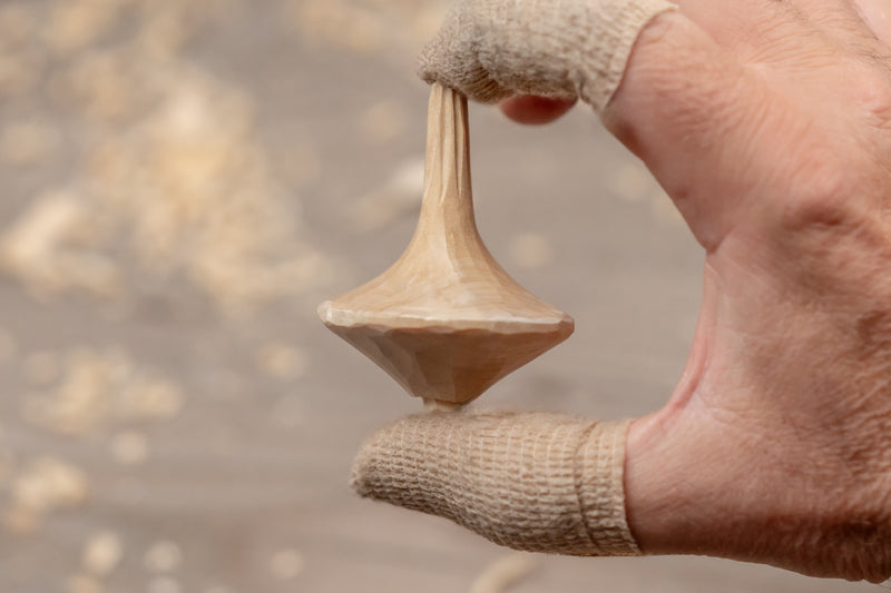 A close-up of a hand wearing beige knit finger gloves holding a small unfinished wooden spinning top against a blurred sandy background.