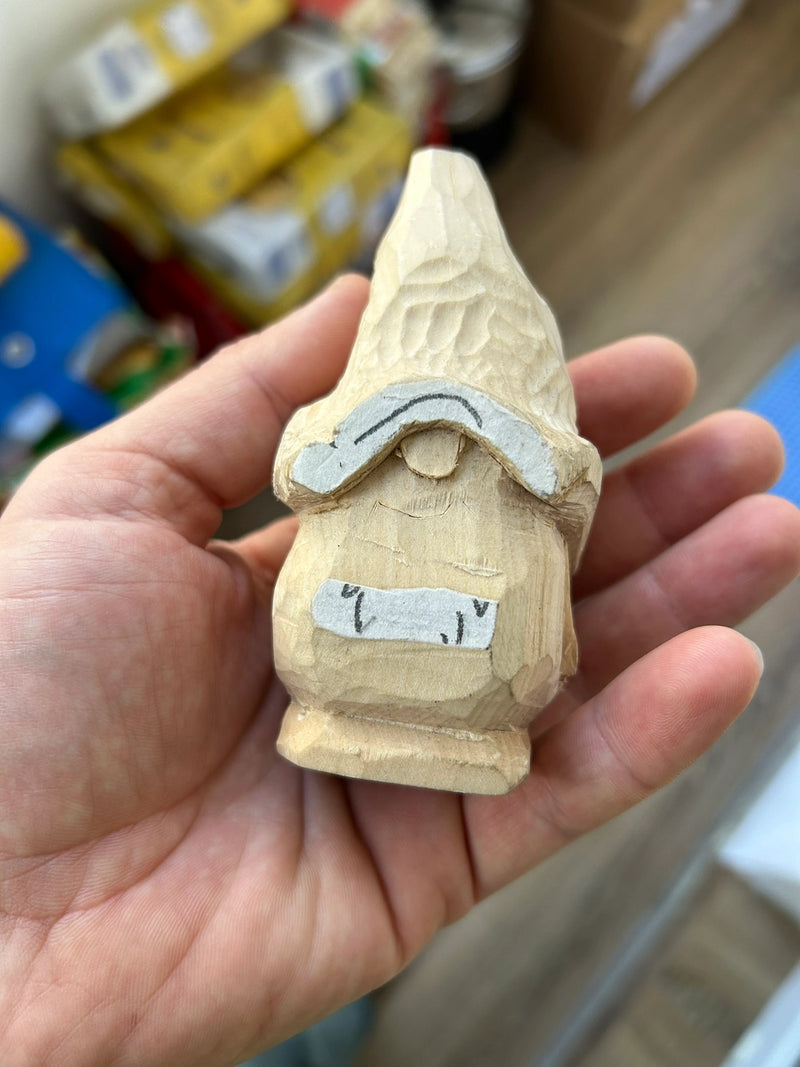 A hand holds a small, hand-carved wooden gnome figurine with a pointed hat and a gray painted roof. The background is a blurry workshop or storage room setting.